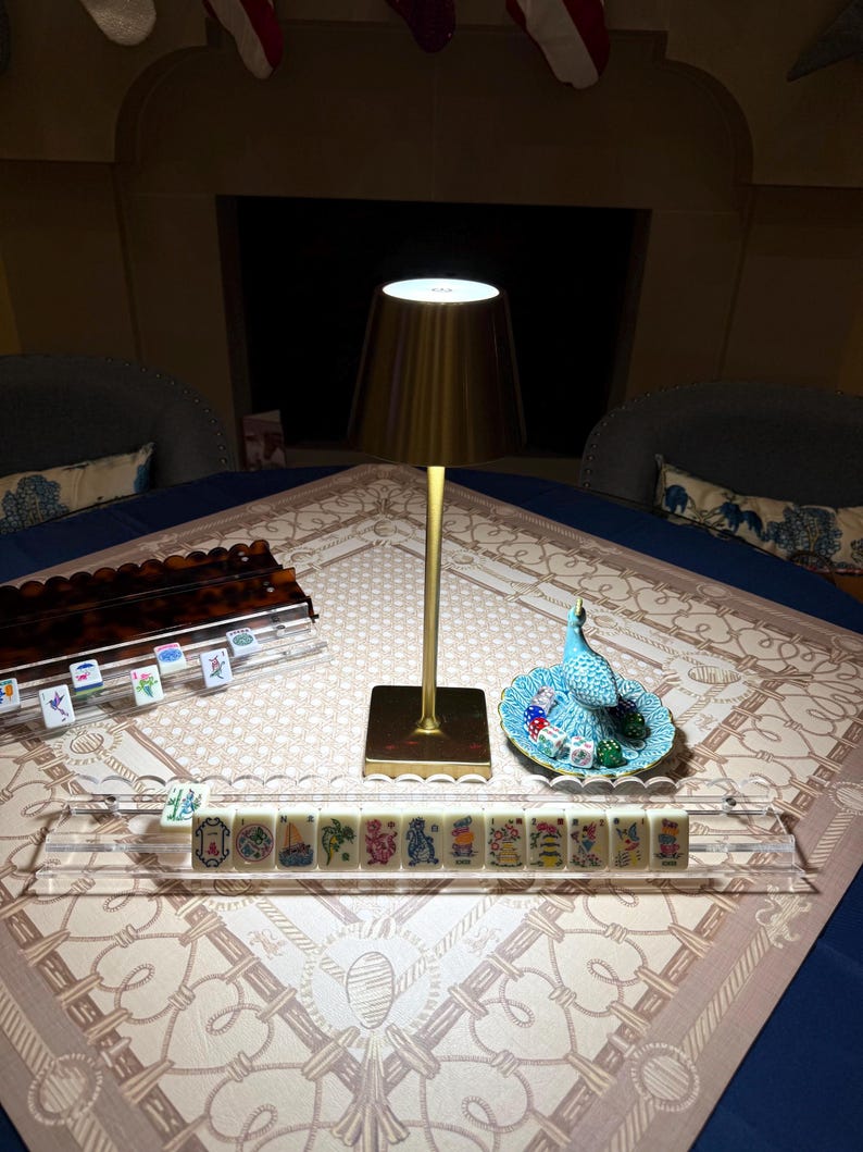May include: A gold-toned metal table lamp with a conical shade stands next to a clear acrylic rack holding mahjong tiles. A small turquoise ceramic peacock figurine sits on a matching scalloped dish containing several colorful dice.