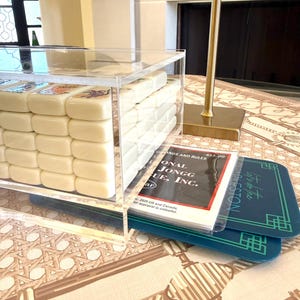 May include: A clear acrylic box containing a set of off-white Mahjong tiles with colorful engraved designs. Beside the box lies a booklet titled National Mahjong League, Inc. Standards and Rules and two dark teal rectangular game score cards.