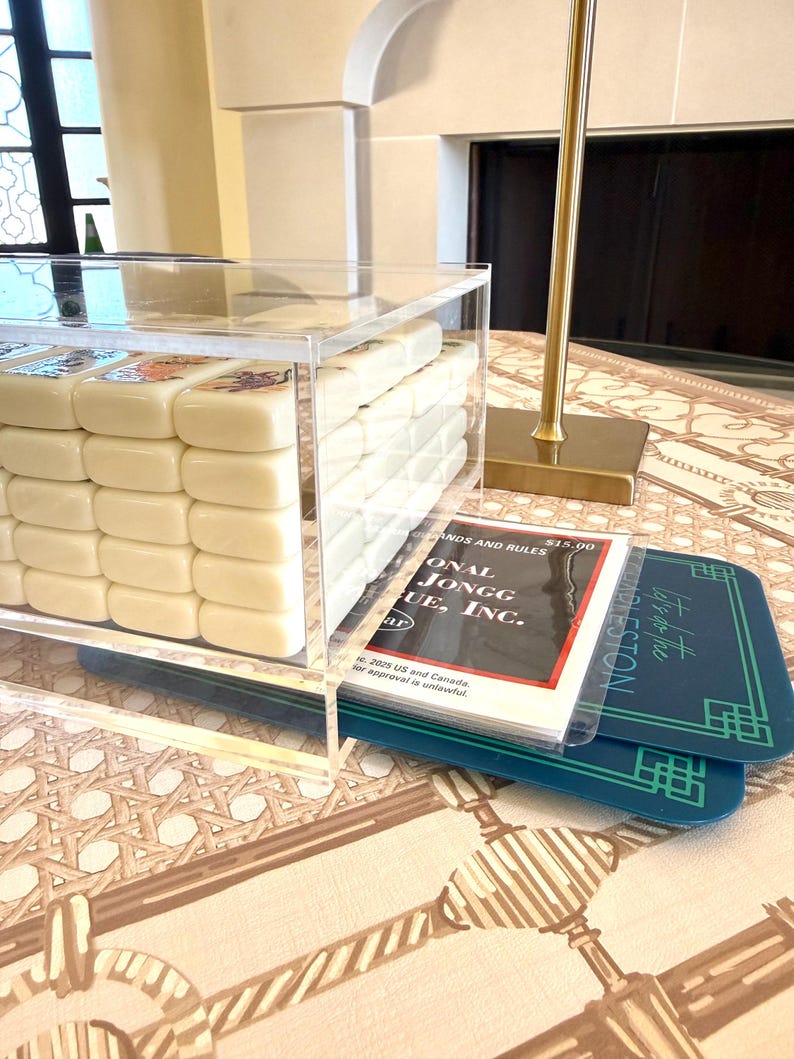 May include: A clear acrylic box containing a set of off-white Mahjong tiles with colorful engraved designs. Beside the box lies a booklet titled National Mahjong League, Inc. Standards and Rules and two dark teal rectangular game score cards.