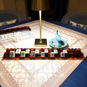 May include: A gold-toned metal table lamp with a conical shade stands beside a tortoise-shell patterned mahjong rack holding white tiles with colorful illustrations. A small turquoise ceramic peacock figurine sits on a matching scalloped dish.