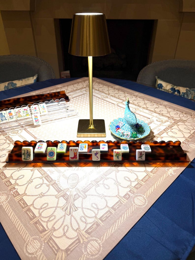 May include: A gold-toned metal table lamp with a conical shade stands beside a tortoise-shell patterned mahjong rack holding white tiles with colorful illustrations. A small turquoise ceramic peacock figurine sits on a matching scalloped dish.
