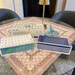May include: Three sets of Mahjong tiles in clear acrylic cases are displayed on a patterned tablecloth. The tiles have various designs and colors. A small gold lamp and a decorative peacock figurine are also on the table. The table is made of glass.