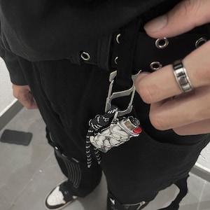 Puede incluir: Una funda de metal plateado para encendedor con un dise&ntilde;o intrincado, org&aacute;nico y calado tipo red, sujeta a un cintur&oacute;n negro mediante un mosquet&oacute;n plateado y un cord&oacute;n de paracord negro y blanco, que sostiene un encendedor blanco est&aacute;ndar.
