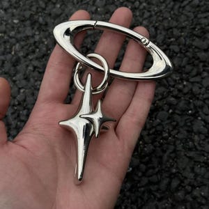 May include: A polished silver-tone metal keyring featuring a large oval carabiner clip attached to a smaller circular ring. A dimensional, four-pointed star charm with a smaller secondary star overlay is suspended from the circular ring.