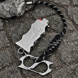 Puede incluir: Una funda de metal plateado para encendedor con un emblema de araña grabado, unida a un cordón de paracord negro y blanco con un mosquetón de metal plateado. La funda tiene una forma contorneada y un botón de encendido rojo en la parte superior.