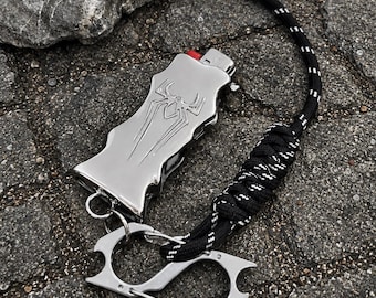 Puede incluir: Una funda de metal plateado para encendedor con un emblema de araña en relieve en la parte frontal. La funda está unida a un cordón de paracord negro y blanco con una sección trenzada y un mosquetón de metal plateado con abrebotellas integrados.