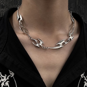 May include: A silver-toned metal chain necklace featuring a series of interconnected, sharp, flame-like or tribal-inspired abstract links. The polished metallic segments are joined by small circular jump rings, creating a continuous collar design.