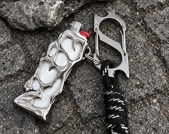 Puede incluir: Un encendedor blanco dentro de una estructura de metal plateado pulido con un diseño orgánico y fluido. El encendedor está unido a un mosquetón plateado en forma de S con abridor de botellas y un cordón negro con detalles tejidos en blanco.
