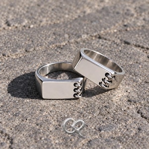 May include: Two polished silver-toned metal rings featuring a rectangular flat face and a side detail resembling a spiral notebook binding with three small metallic loops. The rings have a smooth, reflective surface and a wide, solid band construction.