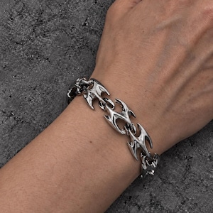 Puede incluir: Una pulsera de metal plateado que presenta una serie de eslabones interconectados con formas afiladas, similares a llamas o alas. La superficie pulida y reflectante de los eslabones crea un patrón distintivo y repetitivo alrededor de la muñeca.