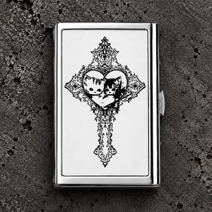 May include: A rectangular silver metal cigarette case featuring a black engraved design of two kittens nestled inside a heart, which is centred within an ornate, cross-shaped filigree pattern. The case has a polished, reflective finish.