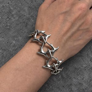 May include: A silver-toned metal bracelet composed of interconnected, thorn-like links. Each link features a sharp, multi-pointed design with a polished, reflective surface. The bracelet wraps around the wrist, showcasing a repeating, angular pattern.