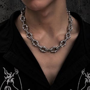 May include: A silver-toned chain necklace featuring oval links with protruding, star-like spikes at each connection point. The necklace rests around the neck, showcasing a metallic finish and a repeating geometric design throughout the entire length of the chain.