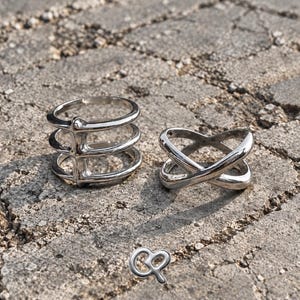 May include: Two silver-toned rings. One ring features three parallel bands connected by a vertical bar. The second ring consists of two bands that cross over each other in an X-shape. A small silver-toned logo with the letters CP is visible below.