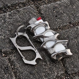 May include: A white lighter encased in a polished, silver-toned metal sleeve with an organic, fluid, and spiked design. A matching silver-toned metal carabiner with a spring-loaded gate is attached to the lighter sleeve by a circular metal ring.