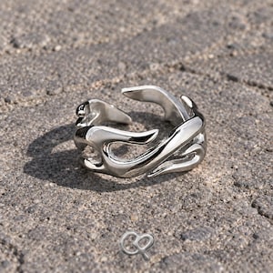 Puede incluir: Un anillo de tono plateado pulido con un diseño abierto y fluido que presenta formas curvas similares a llamas que envuelven el dedo. El metal tiene una superficie lisa y reflectante con una abertura ajustable en la parte superior del diseño.