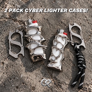 Puede incluir: Dos encendedores blancos dentro de fundas de metal plateado con formas orgánicas y el texto 2 PACK CYBER LIGHTER CASES! visible. Cada funda tiene un mosquetón de metal y una incluye un cordón con patrón en blanco y negro.