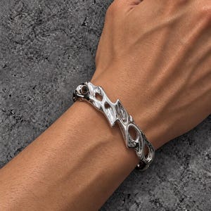 May include: A silver-toned metal cuff bracelet featuring an irregular, organic, molten-like surface with several cut-out openings throughout the band. The bracelet has a high-polish, reflective finish and a fluid, asymmetrical design.