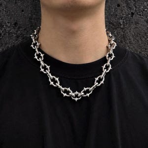 Op de afbeelding: Een zilverkleurige ketting bestaande uit een reeks onderling verbonden ovale schakels. Elke schakel is versierd met vier kleine, puntige stekels die naar buiten steken, wat zorgt voor een getextureerd, doornachtig uiterlijk rondom de halsketting.