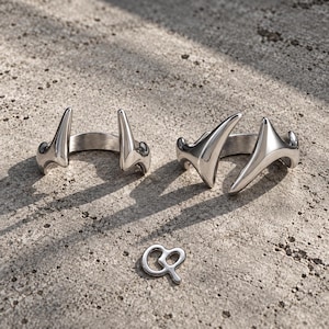 May include: Two silver-toned metal rings featuring a pointed, horn-like design with an open band structure. Below the rings lies a small, silver-toned metal charm shaped like the letters QP, featuring a smooth, reflective, and polished finish.