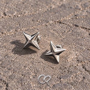 May include: A pair of polished silver-toned stud earrings shaped like four-pointed stars with sharp, defined edges. Each earring features a central raised ridge and a post back. Below the earrings, a small silver-toned ampersand symbol is visible.