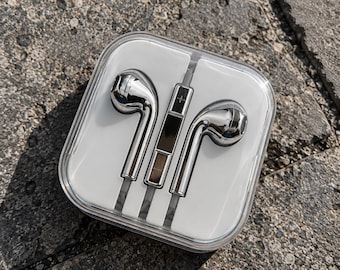 Puede incluir: Un par de auriculares con cable en color plata metálica con un módulo de control central que presenta un signo más, alojados dentro de un estuche protector cuadrado de plástico transparente con un inserto interior blanco que fija el cable.