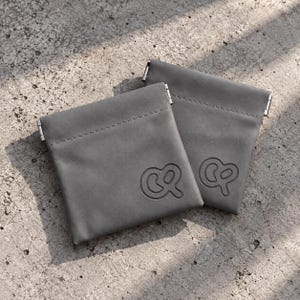 May include: Two square grey pouches made of smooth material, each featuring a debossed pretzel-shaped logo on the front. The pouches have a horizontal stitched seam near the top edge and metal hardware accents on the sides of the opening.