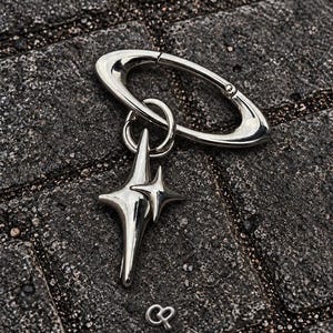 Puede incluir: Un llavero de mosquetón de metal plateado pulido con un dije que presenta dos estrellas de cuatro puntas de diferentes tamaños. El llavero tiene un marco ovalado liso y un pequeño símbolo de logotipo metálico separado con forma de letras CP.