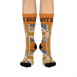 May include: A pair of crew socks featuring a mustard yellow, blue, and white Union Jack flag pattern. The socks are decorated with small white ghost illustrations and white daisy flowers. The text GROOVY BRITAIN is printed in brown at the top.