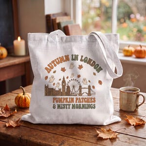 Peut inclure: Un sac fourre-tout en toile blanche avec une impression graphique de la ligne d'horizon de Londres, incluant Big Ben et le London Eye. Le motif comporte des petits fantômes et des feuilles d'automne, avec le texte "AUTUMN IN LONDON PUMPKIN PATCHES & MISTY MORNINGS" imprimé en orange et vert.
