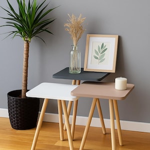 Puede incluir: Un conjunto de tres mesas auxiliares cuadradas en blanco, gris y topo. Cada mesa tiene un marco de madera con patas anguladas. Una impresión botánica enmarcada, una vela y un jarrón de vidrio con flores secas se exhiben sobre las mesas. Una planta en maceta está en el fondo.