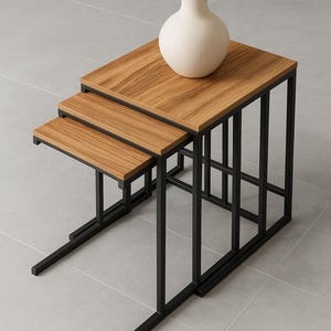 May include: Three modern nesting tables with wooden tops and black metal frames. A cream vase sits on the largest table. The tables are designed to be stacked for space-saving. The tables are approximately 60cm in length.