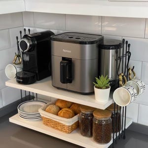 Puede incluir: Una estantería de cocina con una cafetera espresso negra, una freidora de aire plateada y una cafetera. La estantería también contiene tazas blancas, platos, una cesta de panecillos y tarros de granos de café. También hay una pequeña planta suculenta en maceta.