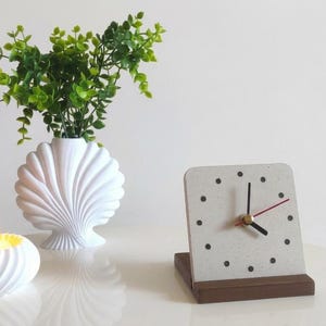 Könnte beinhalten: Eine weiße muschelförmige Vase mit grünem Blattwerk, ein weißer spiralförmiger Kerzenhalter mit brennender Kerze und eine quadratische hellgraue Uhr mit schwarzen Punkt-Stundenmarkierungen auf einem dunkelbraunen Holzsockel.
