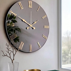 May include: A large circular wall clock with a dark, reflective surface and gold-coloured Roman numerals. The clock features slender gold-coloured hour, minute, and second hands centred on the face. The design is simple, featuring a smooth, dark finish that reflects the surrounding environment, including nearby greenery.