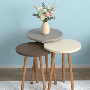 Puede incluir: Un juego de tres mesas nido redondas con superficies lisas en tonos gris, marrón y crema, sostenidas por patas de madera clara. Un pequeño jarrón de cerámica de color claro con un ramo de flores de color rosa pálido descansa sobre la mesa.
