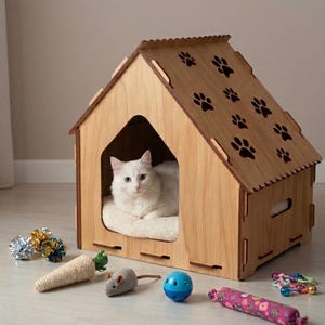 Puede incluir: Una casa para mascotas de madera clara con un techo a dos aguas que presenta diseños de huellas recortados. Un gato blanco descansa en su interior sobre un cojín suave. Frente a la casa hay varios juguetes, incluyendo una zanahoria de sisal.