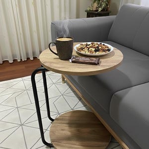May include: A small, round wooden side table with a black metal frame. A cup of coffee, a plate of nuts and dried fruit, and a chocolate bar sit on the table. The table is next to a gray sofa.