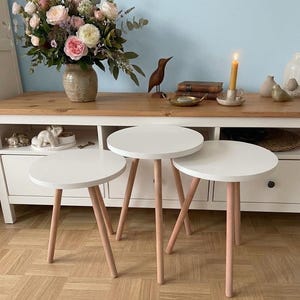 Pode incluir: Três mesas laterais redondas brancas com pernas de madeira rosa claro. As mesas são de tamanhos diferentes e estão dispostas em grupo. Um vaso de flores e outros objetos decorativos estão no fundo.