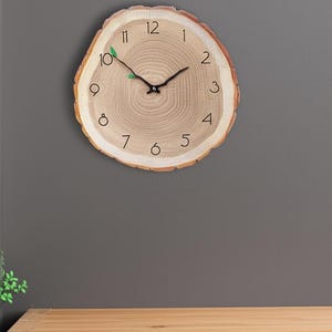 Könnte beinhalten: Eine runde Holzuhr, die einem Baumstammquerschnitt ähnelt und an einer grauen Wand hängt. Das Zifferblatt der Uhr hat schwarze Ziffern und Zeiger, mit einem kleinen grünen Blattdetail bei 10 Uhr. Der Durchmesser der Uhr beträgt ca. 25 cm.
