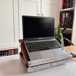 May include: A gray laptop with a black screen and keyboard sits on a wooden laptop stand. The stand features a dark brown wood grain surface and a gray, textured fabric base with a padded front edge to hold the device securely in place.