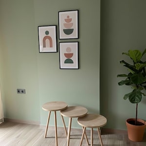 May include: Three small, round wooden side tables with angled legs are arranged in front of a sage green wall. Above the tables, three framed abstract art prints feature geometric shapes in muted colors. A potted plant sits to the right.
