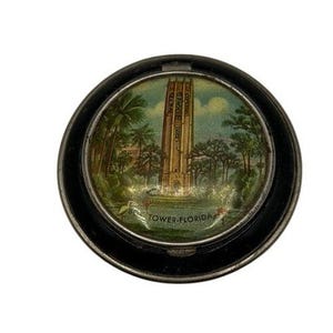 May include: A circular, black metal trinket box featuring a painted landscape with a tall tower, trees, and water. The text "Bok Tower-Florida" is at the bottom. The artwork uses green, brown, and blue hues.