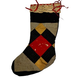 Peut inclure: Une chaussette de Noël faite à la main avec un motif patchwork. Le motif est composé de losanges noirs, rouges et dorés sur un fond beige. Le haut est orné d'une bordure en dentelle crème et de rubans rouges.