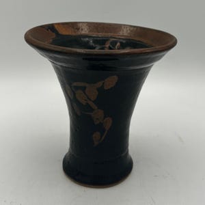 May include: A black ceramic vase with a flared top and a brown rim. The vase features a brown floral design on the side. The vase is approximately 6 inches tall.