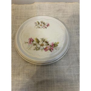 Puede incluir: Caja de baratijas redonda de porcelana blanca con diseño floral. La tapa presenta una delicada disposición de flores rosas y blancas con hojas verdes, sobre un fondo blanco con un borde de encaje. El borde tiene un ribete dorado.