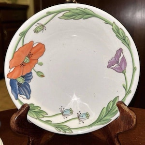 May include: A white ceramic bowl with a green leafy vine border and colorful poppy flower designs. The bowl is on a wooden stand.