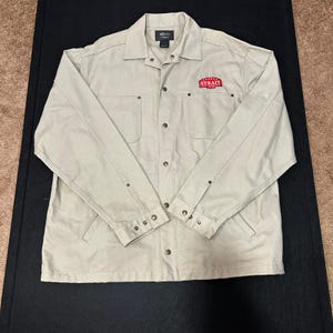 May include: A tan button-up jacket with two chest pockets and a collar. The jacket has a red patch that reads "Straight" in white letters. The jacket has long sleeves and button closures. The jacket is made of a durable fabric.