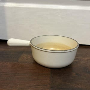 May include: White enameled cast iron saucepan with a single handle. The saucepan is empty and has a light brown interior.