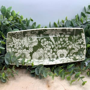 May include: Rectangular ceramic tray with a green background and a white floral pattern. The tray has rounded corners and a slightly raised edge. The floral design includes various flowers and foliage. The tray is surrounded by green leaves.